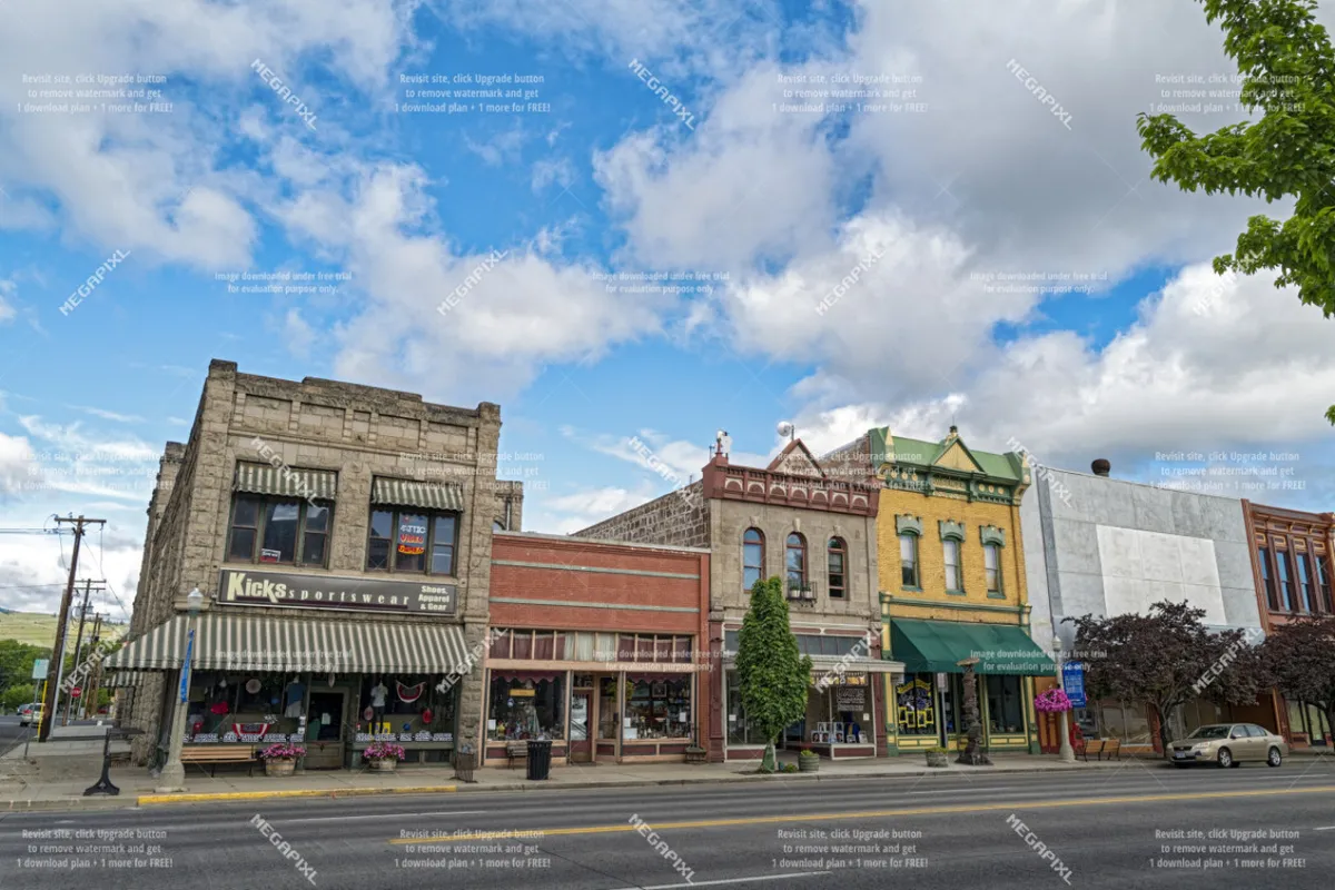 A lively downtown streetscape featuring historic brick buildings, colorful storefronts, and sidewalks adorned with planters. Local shops and cafés suggest a welcoming atmosphere perfect for wandering and discovering Baker City's charm.