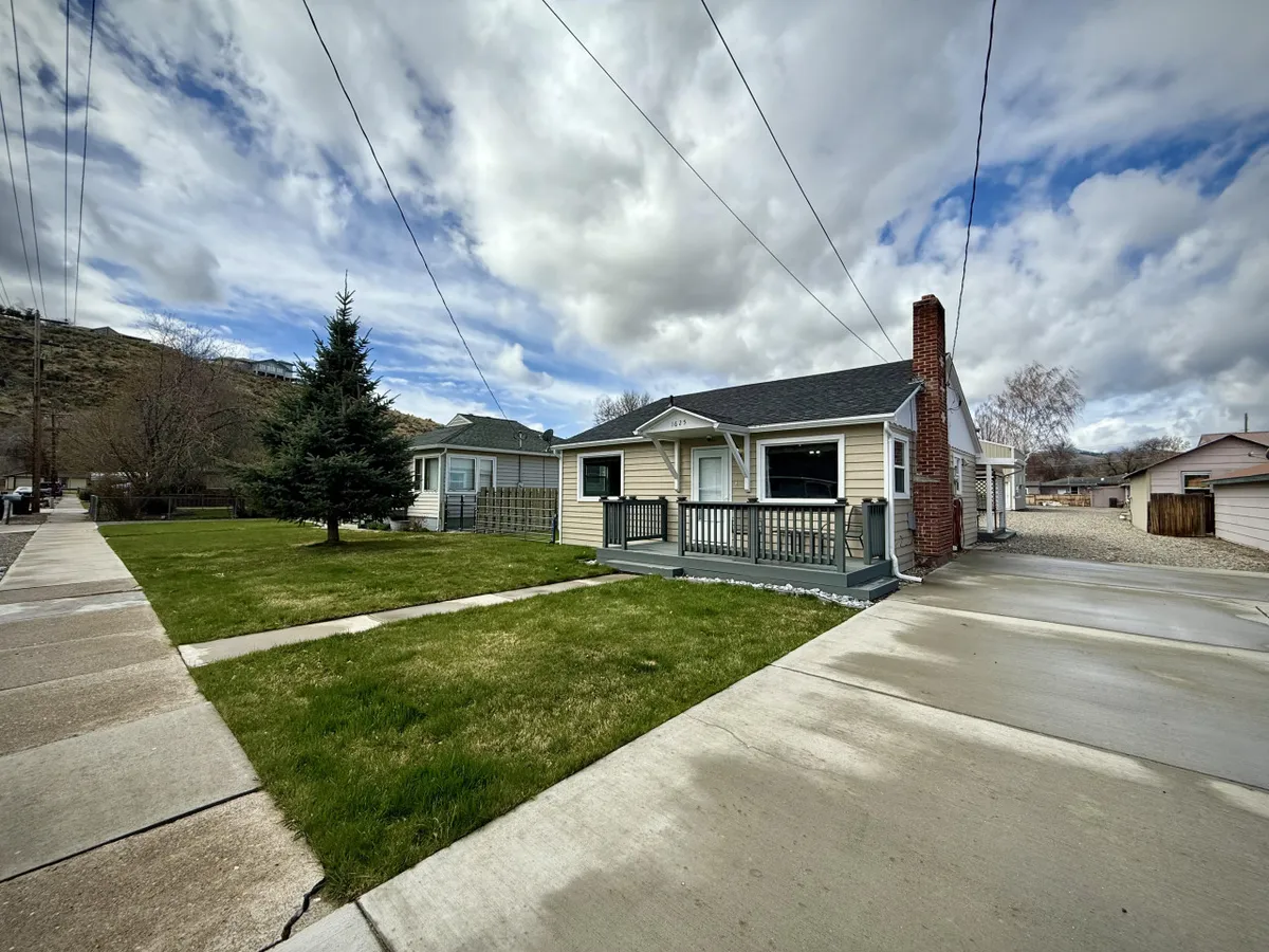 A charming front porch with comfortable seating looks out over a well-maintained lawn and a distinctive brick chimney. The scene is bright, welcoming, and suggests a relaxing place to unwind, with plenty of open space visible for outdoor activities with pets or family.