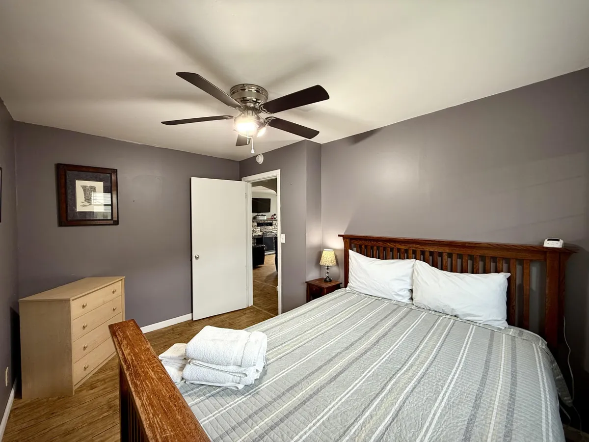 A bright, comfortable bedroom showcasing a queen bed dressed in crisp white linens, a wooden dresser, and a ceiling fan. The window allows in ample natural light, enhancing the room's soothing and airy feel.