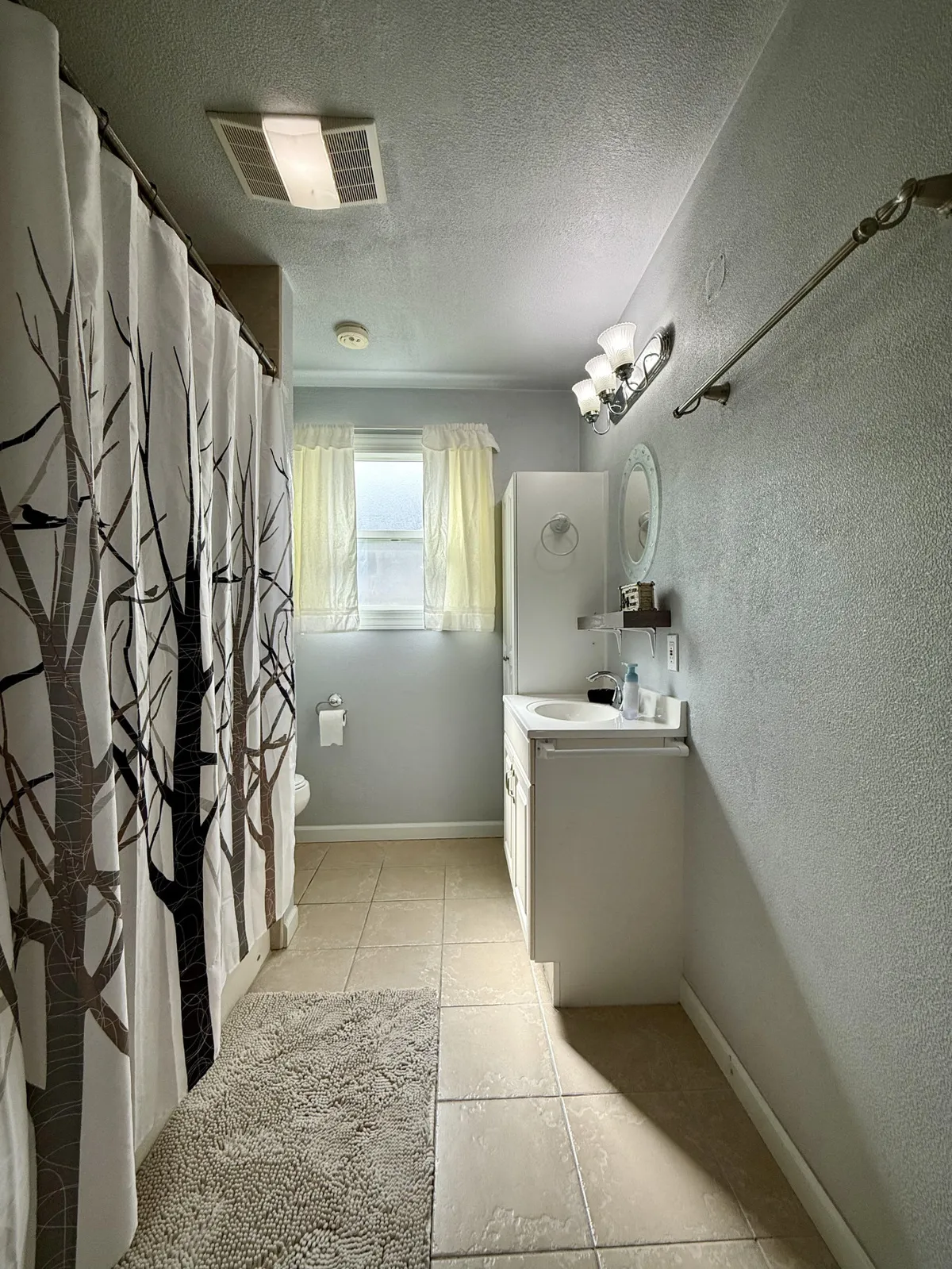A bathroom oasis featuring a full bathtub with a decorative shower curtain, a vanity with a circular mirror, and a window draped with sheer curtains that softly filter the light. The room exudes a sense of calm and rejuvenation, ideal for unwinding.