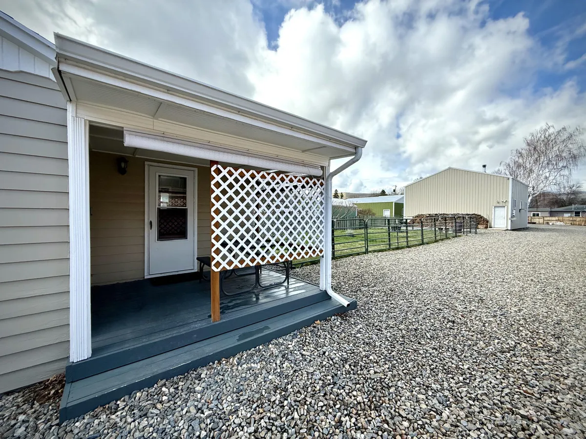 A welcoming backyard scene featuring a covered porch area with a lattice screen, a gravel pathway, and an outbuilding in the background. The spacious yard appears fully fenced and offers plenty of room for pets or people to relax and play. The porch is inviting for both lounging and hosting outdoor gatherings.