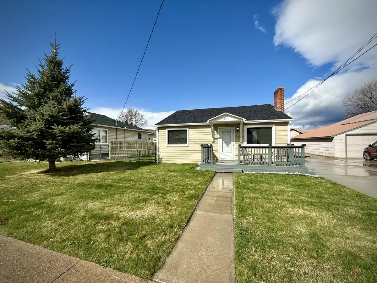 A cheerful exterior view of the yellow house, set amidst a green lawn and shaded by a large pine tree. The front porch features a railing and inviting seating, creating a welcoming entry and a cozy spot to relax outside.