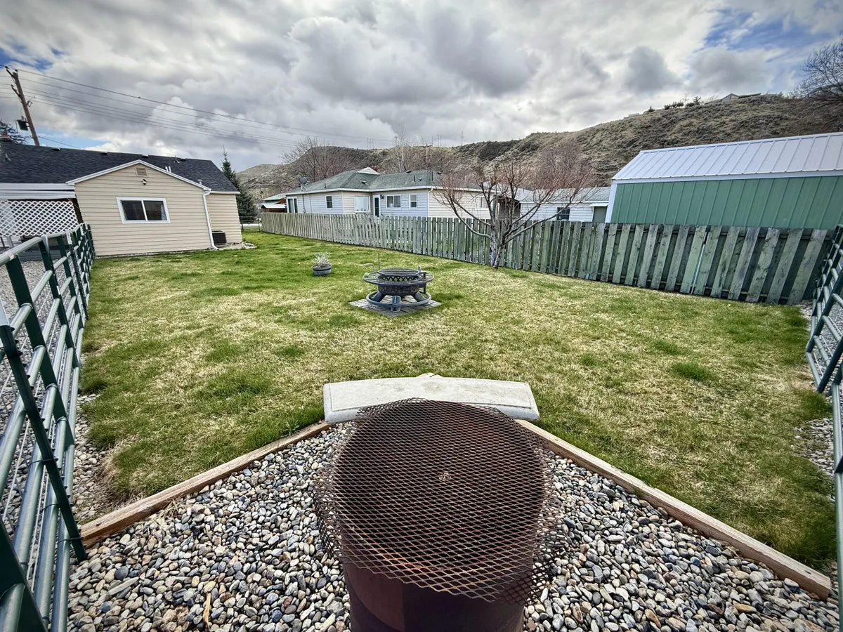 The image focuses on a fire pit surrounded by a grassy area, with views of the fenced yard and neighboring homes. The setting is perfect for evening gatherings, marshmallow roasting, and stargazing beneath the night sky.