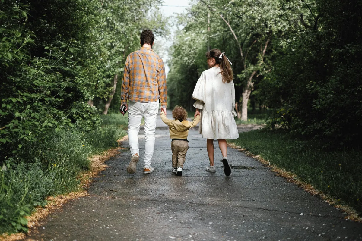 A joyful family walks together along a scenic path surrounded by lush greenery. The child is holding hands with parents, and the natural setting suggests a nearby park or trail—ideal for outdoor recreation and dog walking.