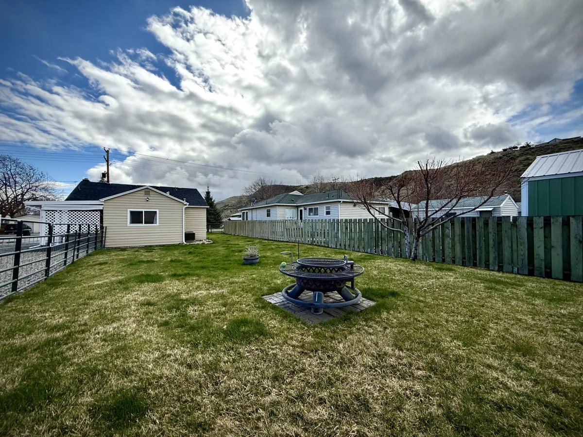 A tranquil backyard with lush green grass, a central fire pit surrounded by seating, and hints of neighboring houses in the background. The sky is partly cloudy, creating a cozy evening atmosphere.