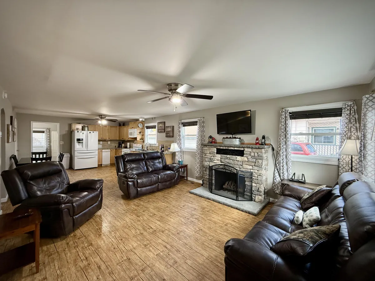 A cozy living room featuring leather sofas arranged around a prominent stone gas fireplace, with a television nearby and an open view to the adjacent kitchen area. The space is inviting for gatherings, relaxation, and entertainment.
