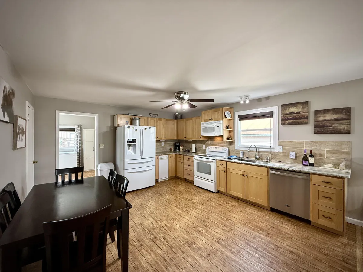 A warm, welcoming kitchen and dining space featuring a central kitchen island, a large dining table with seating for six, stainless steel refrigerator, oven, and natural light streaming in. The setup highlights areas for both cooking and gathering, with modern appliances visible and a comfortable flow between kitchen and dining zones.