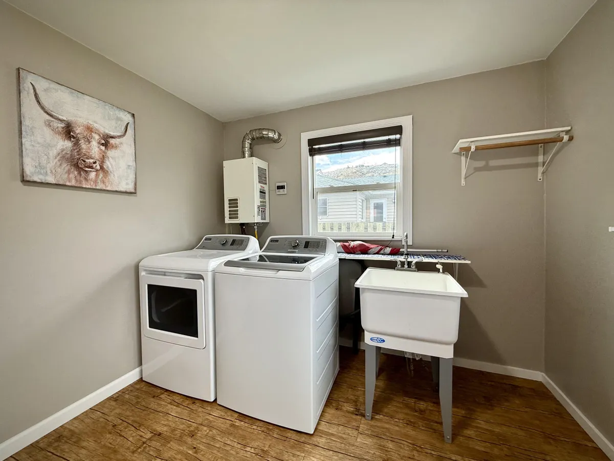 A functional and welcoming laundry room containing a washing machine and dryer, a utility sink, a window with a pleasant view, and a whimsical animal portrait. The space is practical yet bright, making laundry tasks enjoyable.